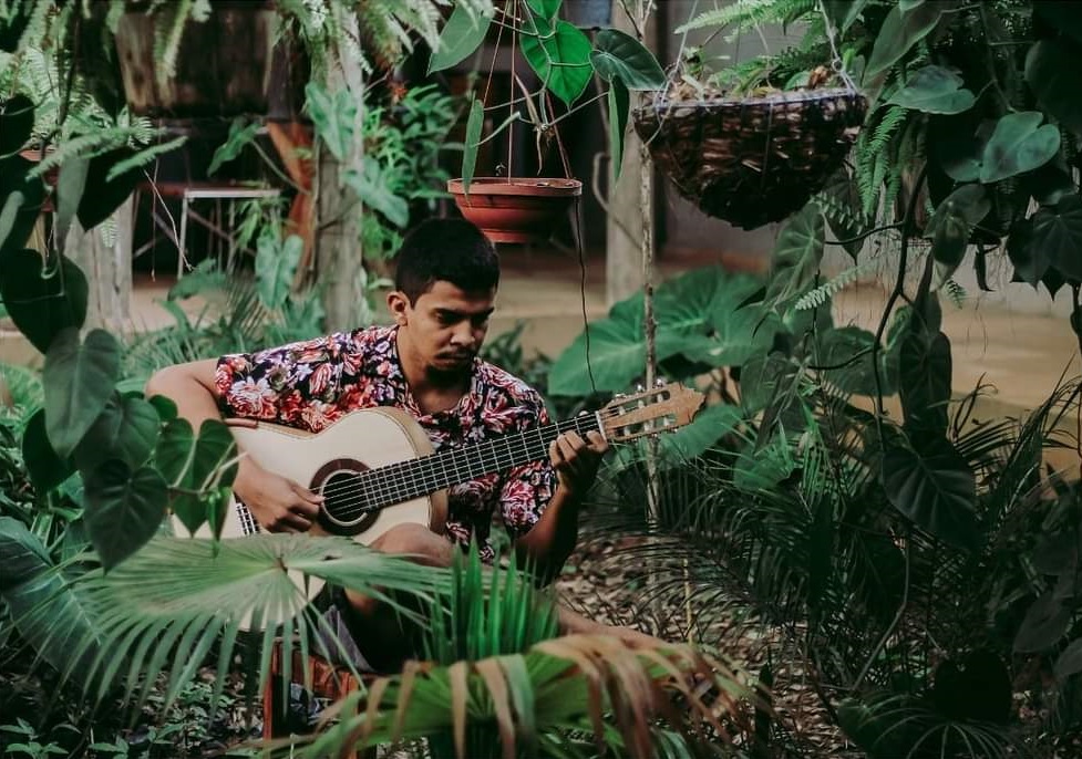Edésio César tocando violão entre plantas, em um registro de sua trajetória musical